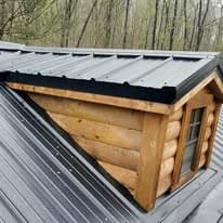 Log cabin roof with metal covering and skylight, surrounded by dense forest.