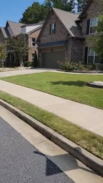 Residential street view featuring a stone house, manicured lawn, and paved sidewalk.